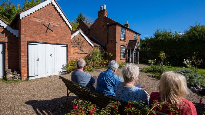 Group of four elderly visitors sitting on a bench in the garden at The Firs, facing the house